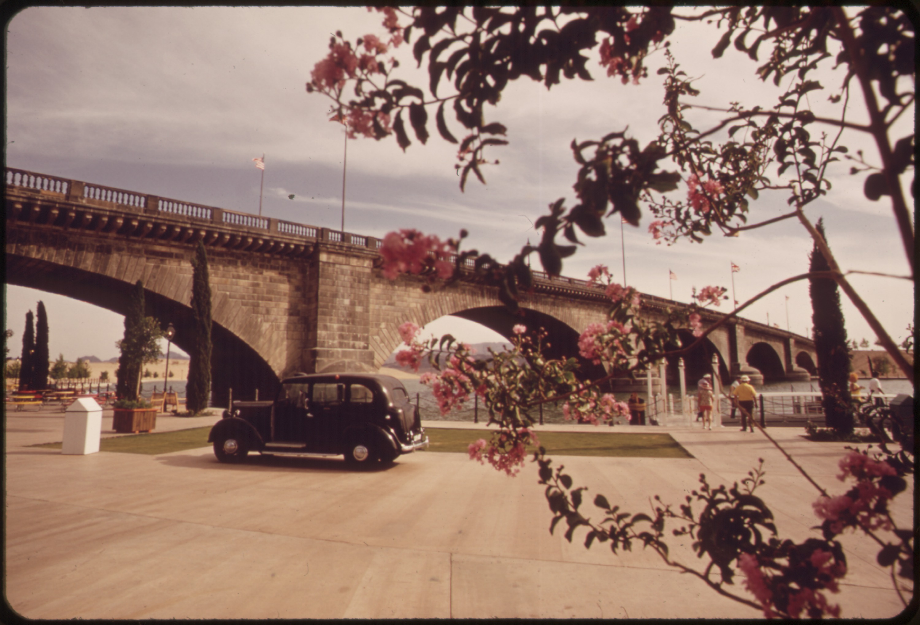 An archival view of London Bridge’s stone arches in Lake Havasu City, with flowers in the foreground and a black car beside the channel.