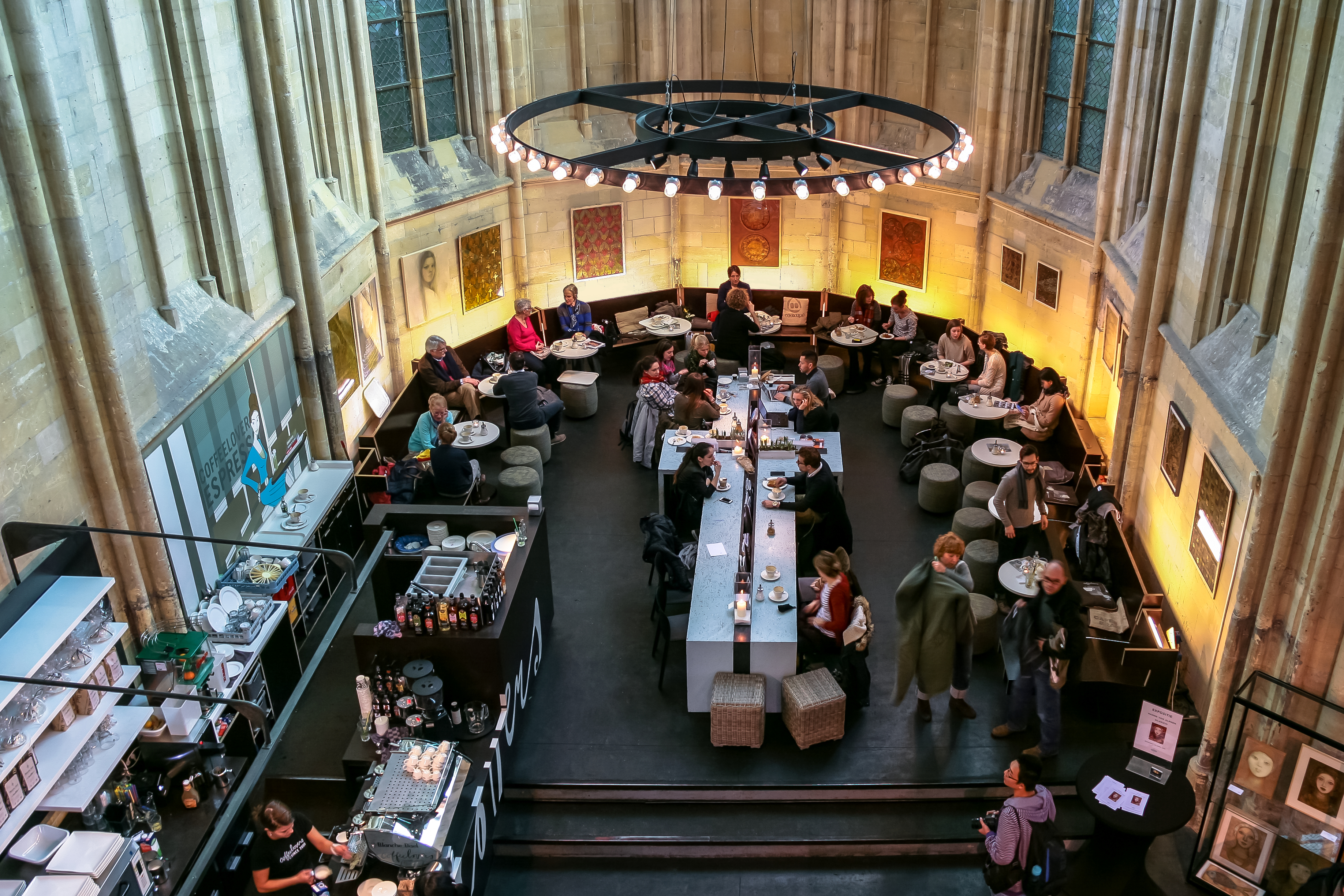 An elevated view of customers at Coffeelovers café beneath the former choir’s tall stone windows, with a cross-shaped central table and a circular hanging light.