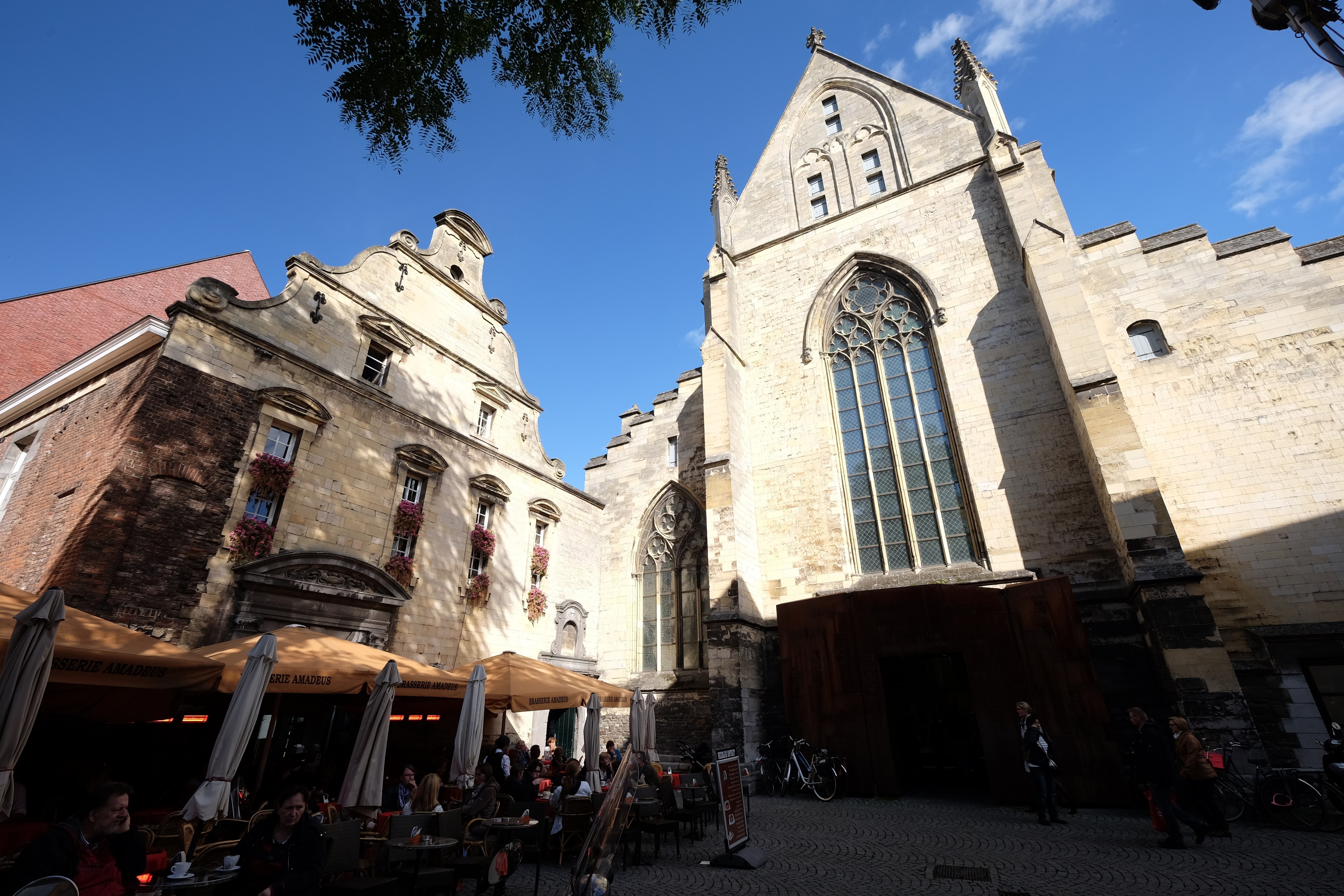 The pale stone Gothic façade of the former Dominican church in Maastricht, with a tall pointed window, neighboring buildings and café terraces in front.