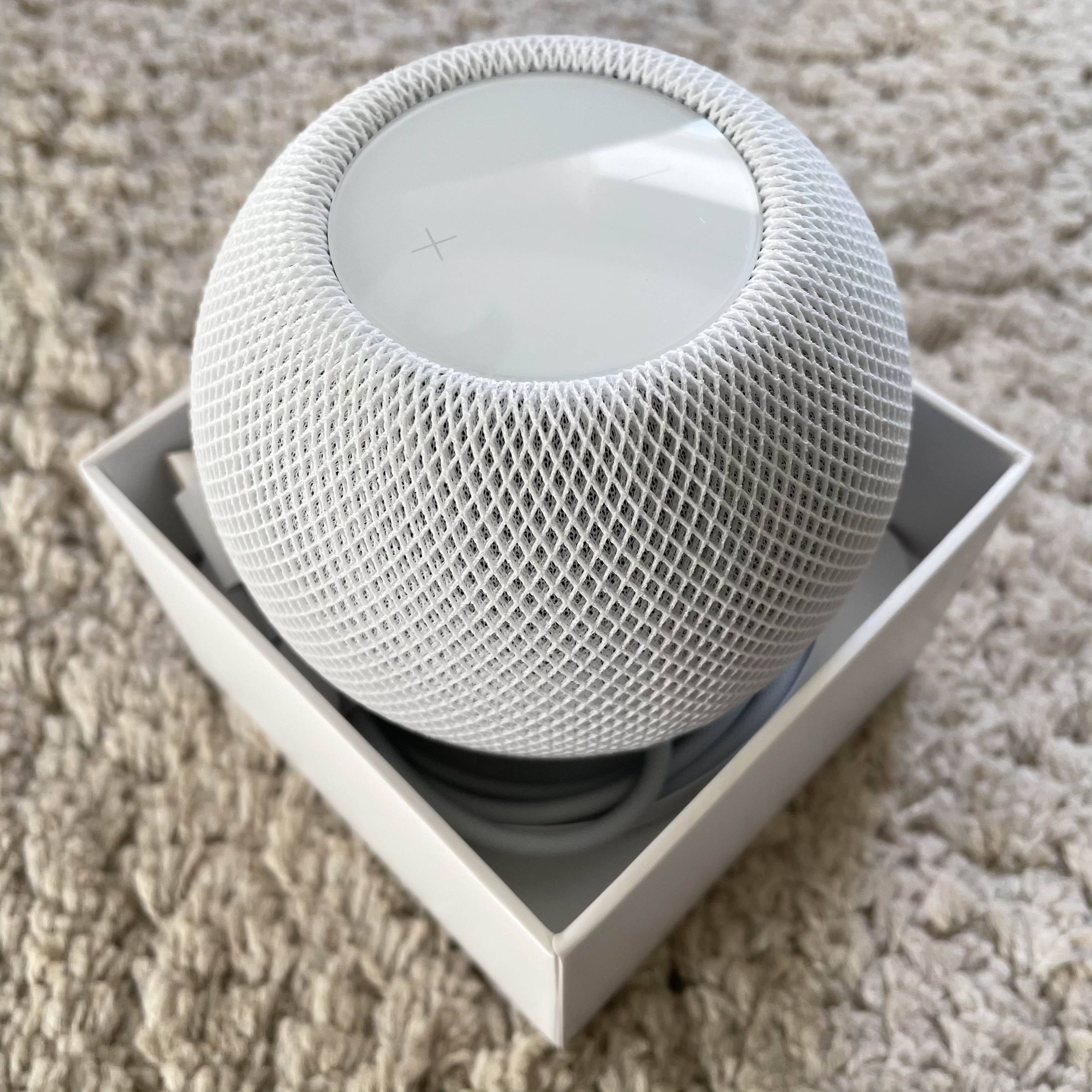 A white original HomePod mini sitting in its open packaging on a textured beige surface.