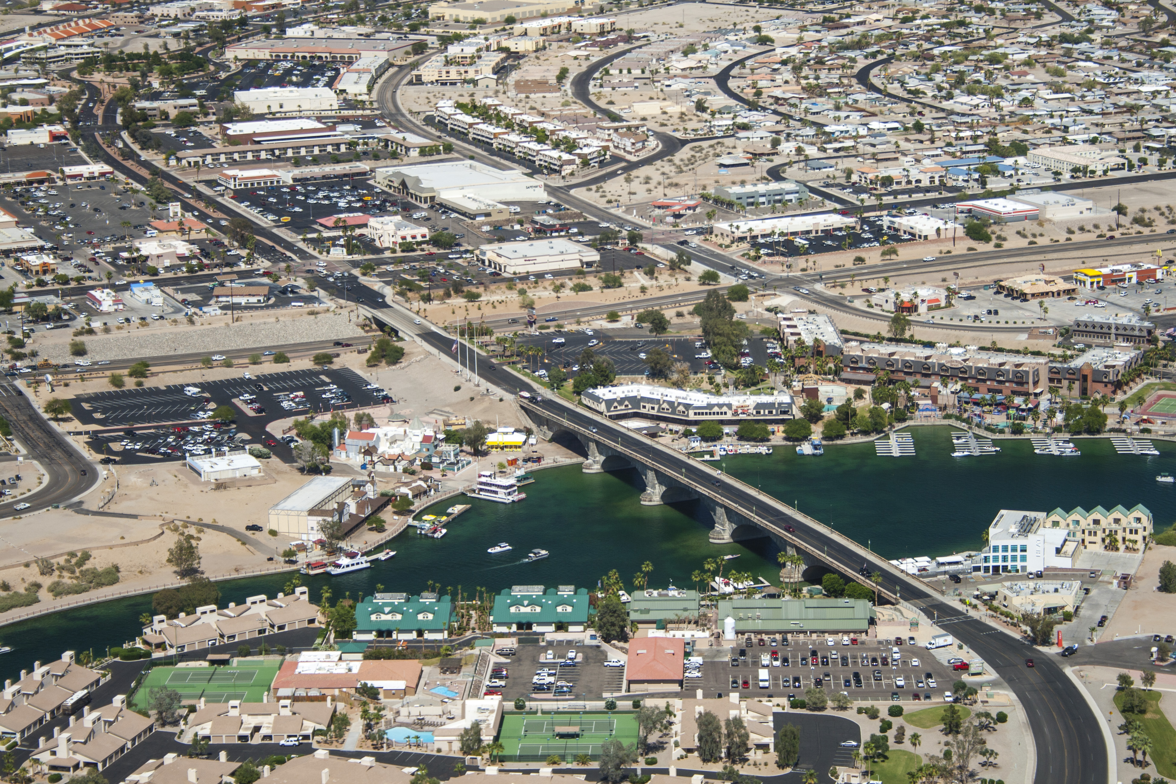 An aerial view of London Bridge crossing a blue-green channel in Lake Havasu City, surrounded by roads, waterfront buildings and desert development.
